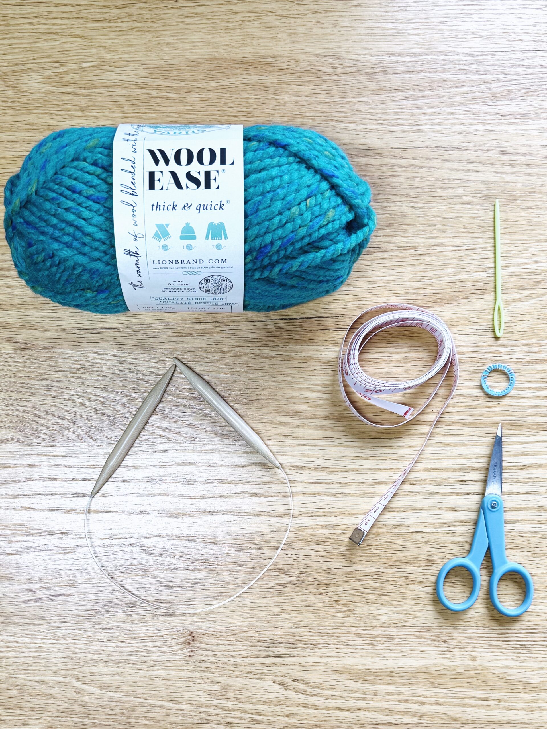 Kids' first knitting supplies for lessons laid on table: Wool-Ease Thick And Quick super-bulky yarn in "Lagoon" blue, bamboo knitting needles US No 13 (9mm), soft measuring tape, darning needle, stitch marker, and sewing scissors. 