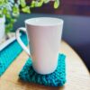 A white coffee cup sits on top of a beginner knit coaster knit with blue super-bulky yarn on a wooden table near a window and potted plant.