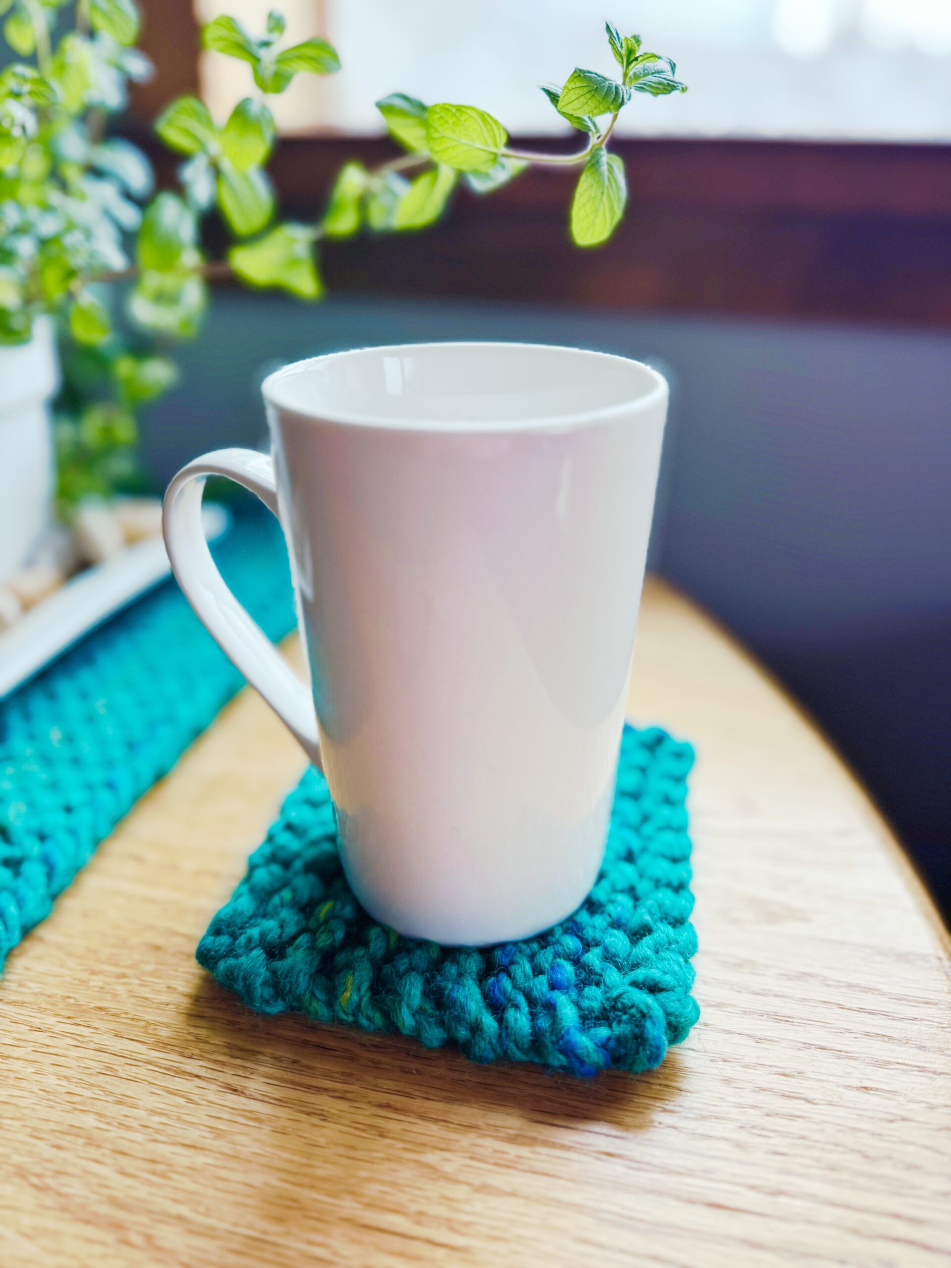 A white coffee cup sits on top of a beginner knit coaster knit with blue super-bulky yarn on a wooden table near a window and potted plant.