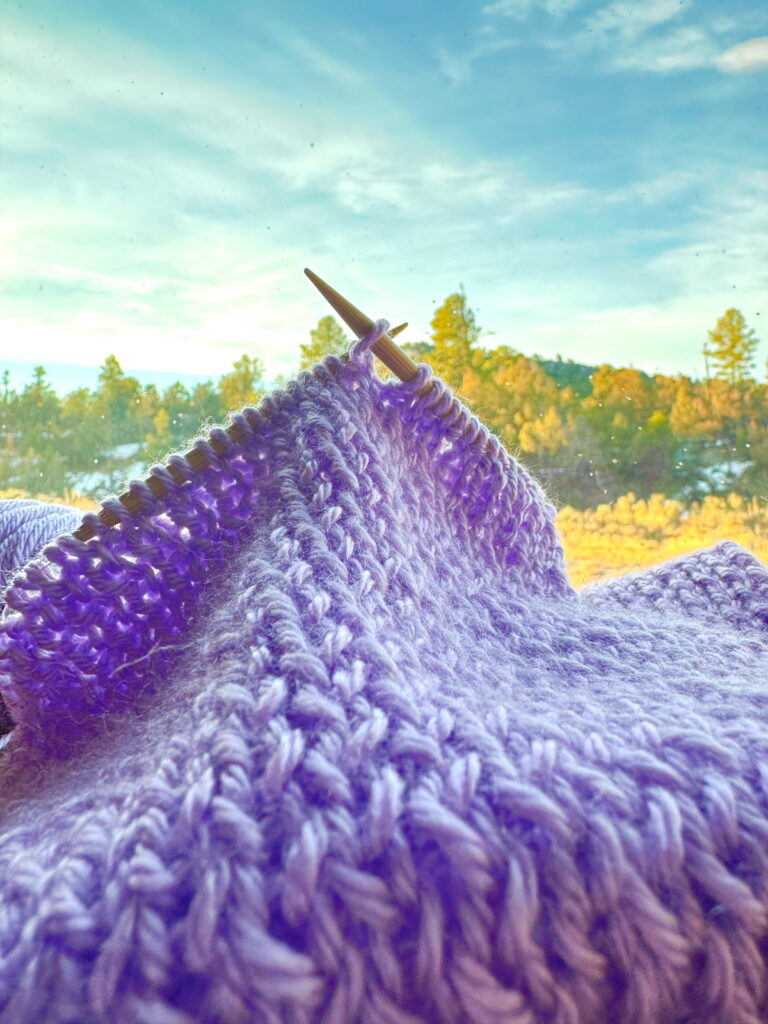 A knit scarf on the windowsill in front of mountain landscape shows how uneven tension can look like a mistake that needs to be ripped out. It does not: just block when finished.