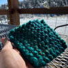 A finished super-bulky knit coaster in teal tones held in a hand in front of a purple porch table and snowy mountain scene.
