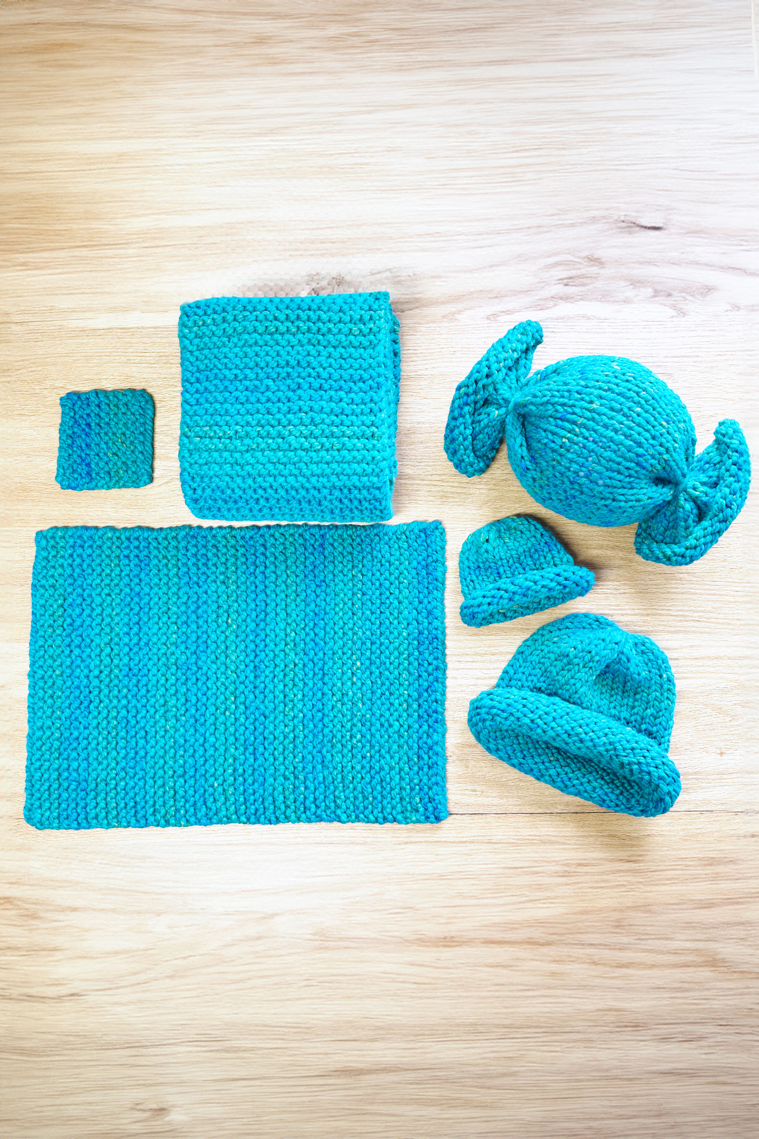 Kids' knitting projects on wooden table - coaster, placemat, scarf, hats, and a candy toy knitted in blue and teal super-bulky yarn.