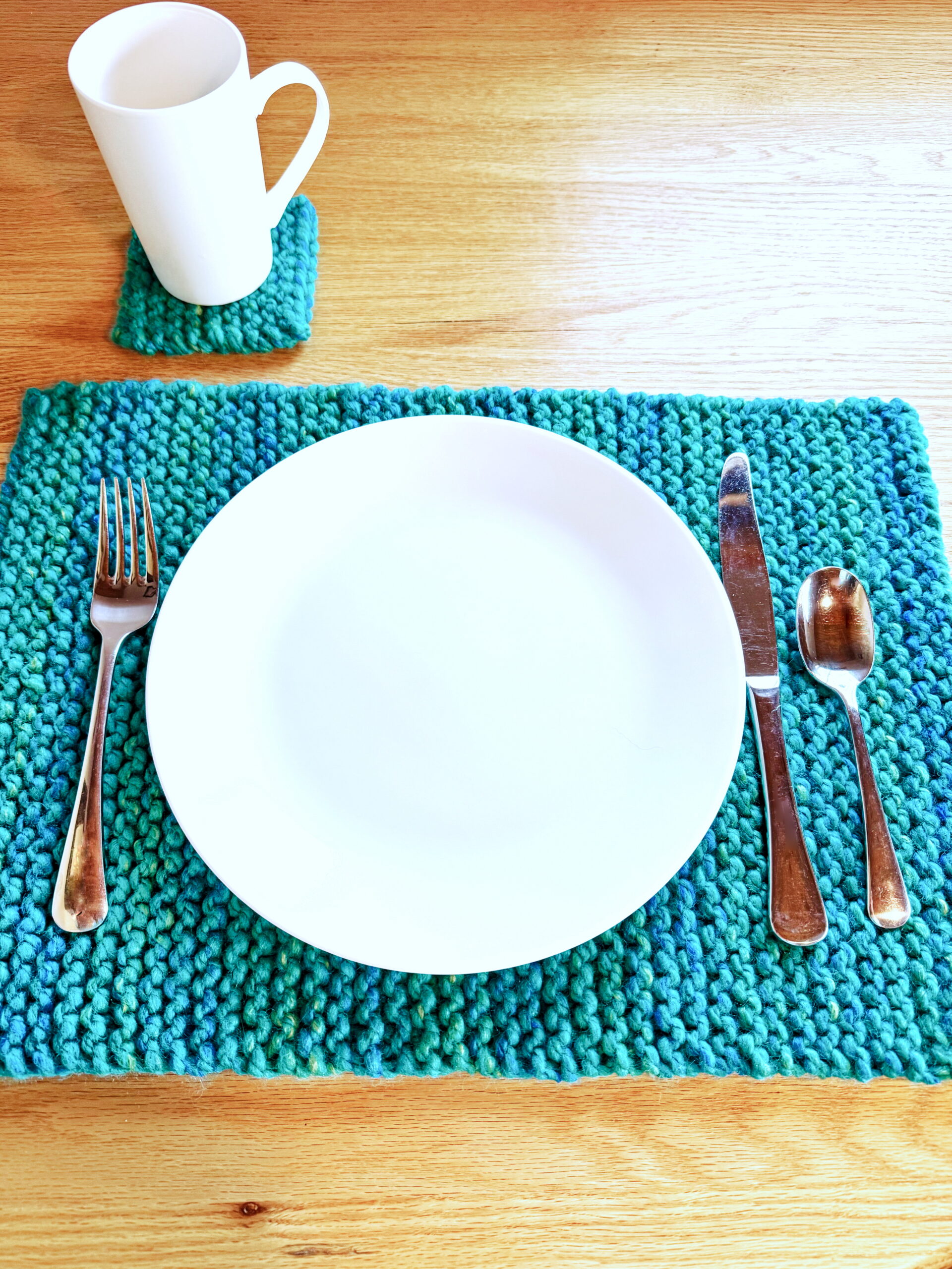 Super-Bulky knit placemat on wooden table with white plate and silverware next to matching knit coaster and mug.
