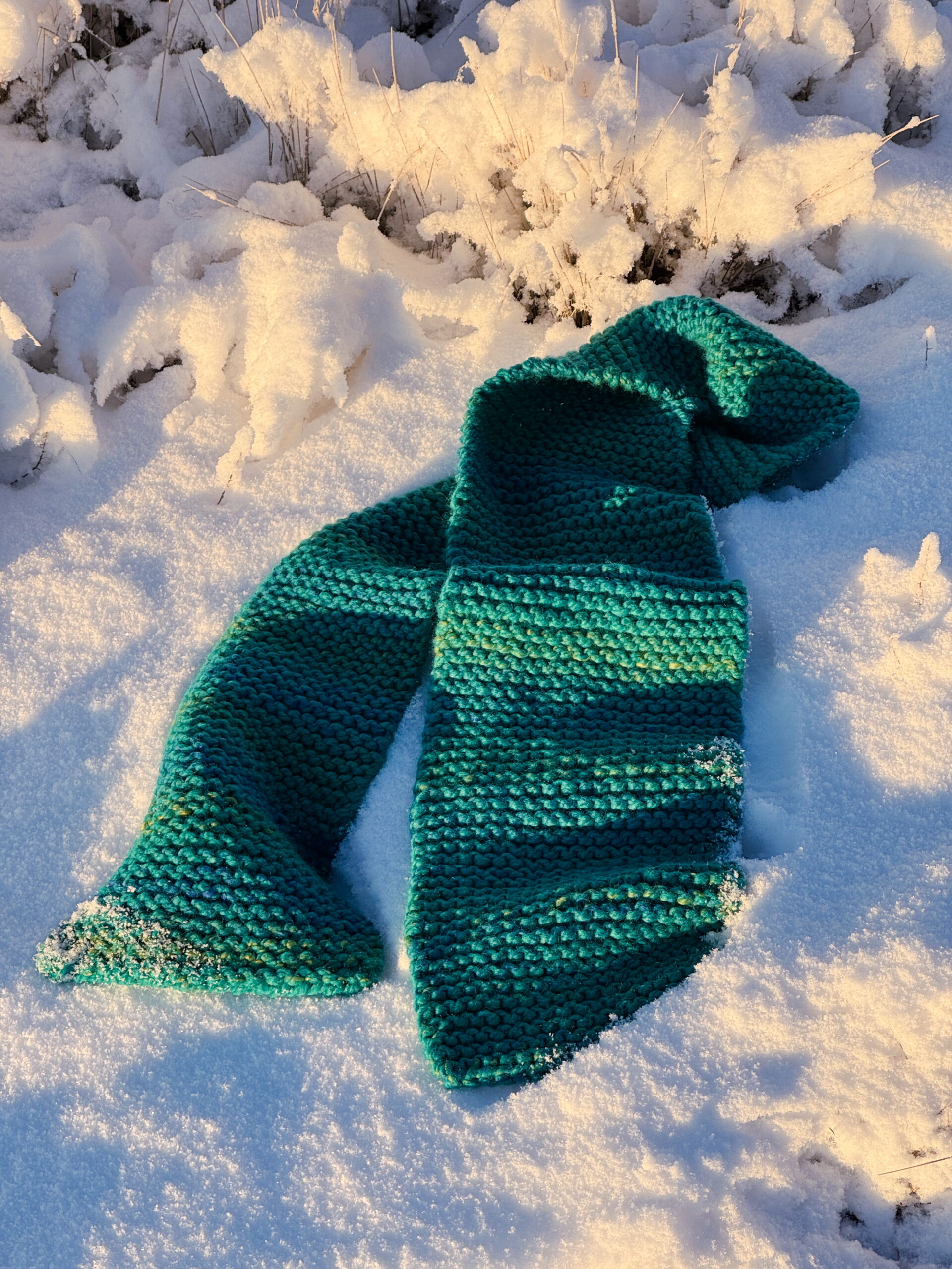 A teal super bulky knit scarf laid haphazardly in fresh snow.