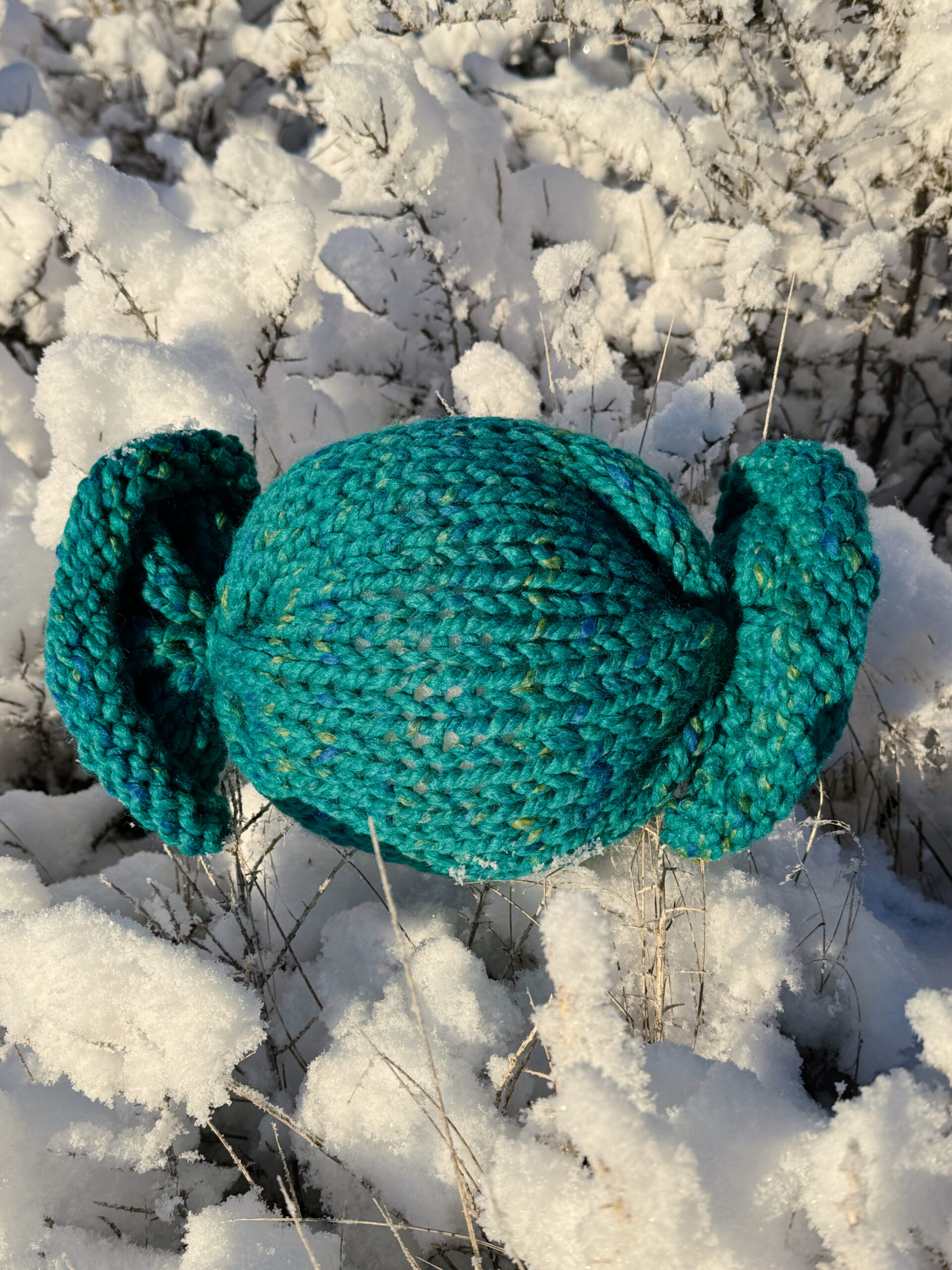 A blue knit candy toy made in the color "Bluegrass" sits on fresh snow.