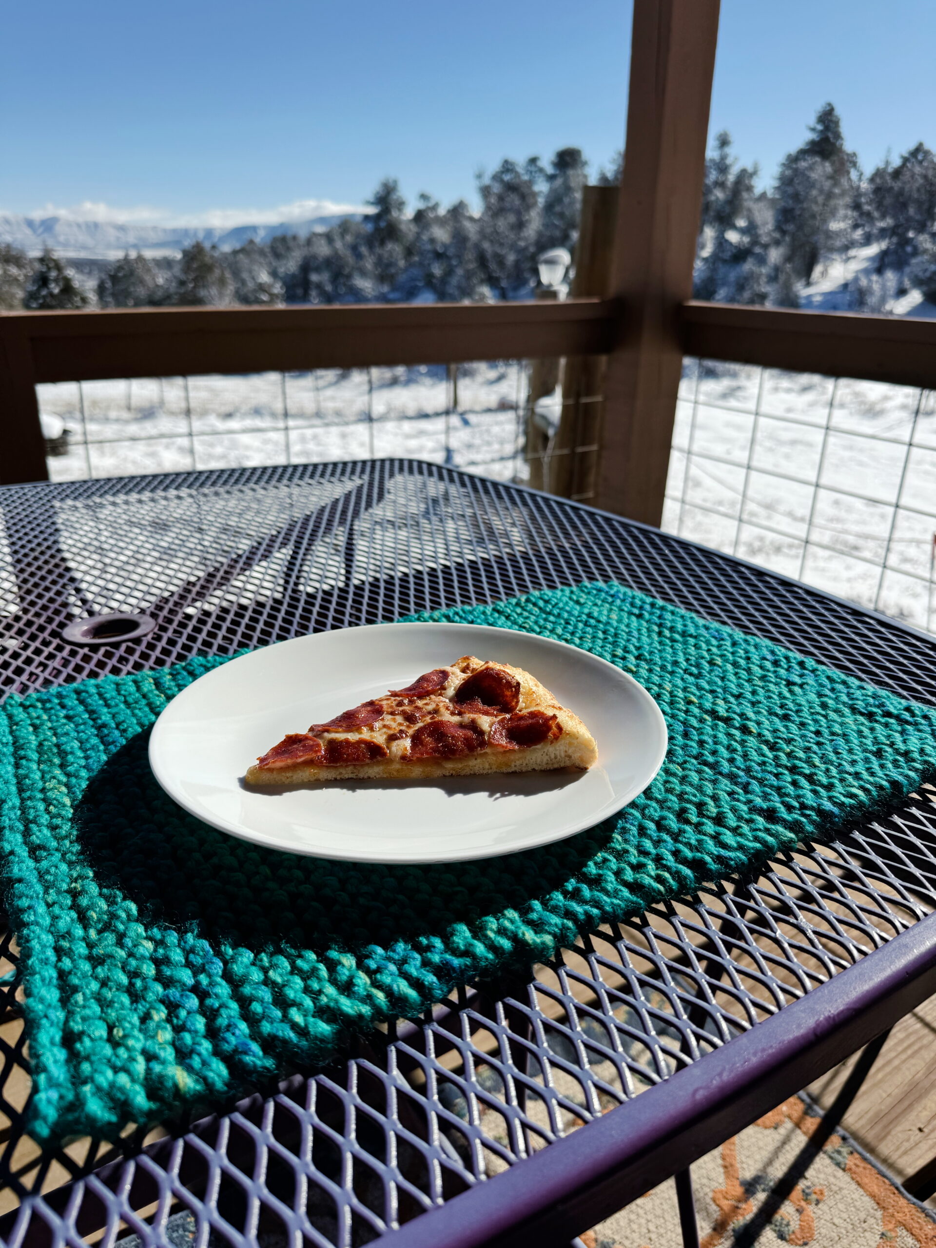 A piece of pizza sits on a white plate on top of the kids' knit placemat in teal on a purple table.