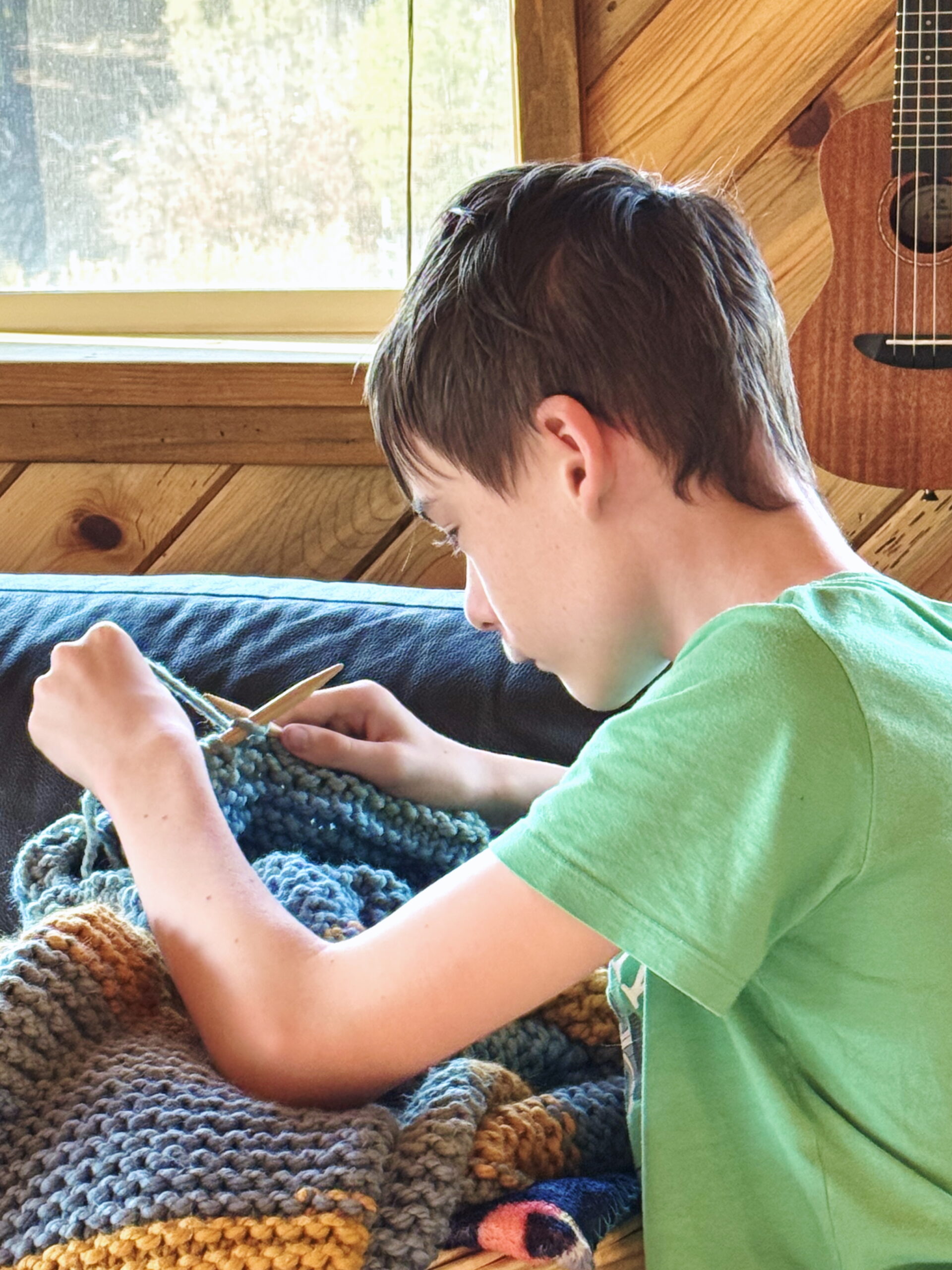A boy knitting by the window learning to knit with super-bulky yarn with Liz Chandler's PurlsAndPixels Knitting Lessons.