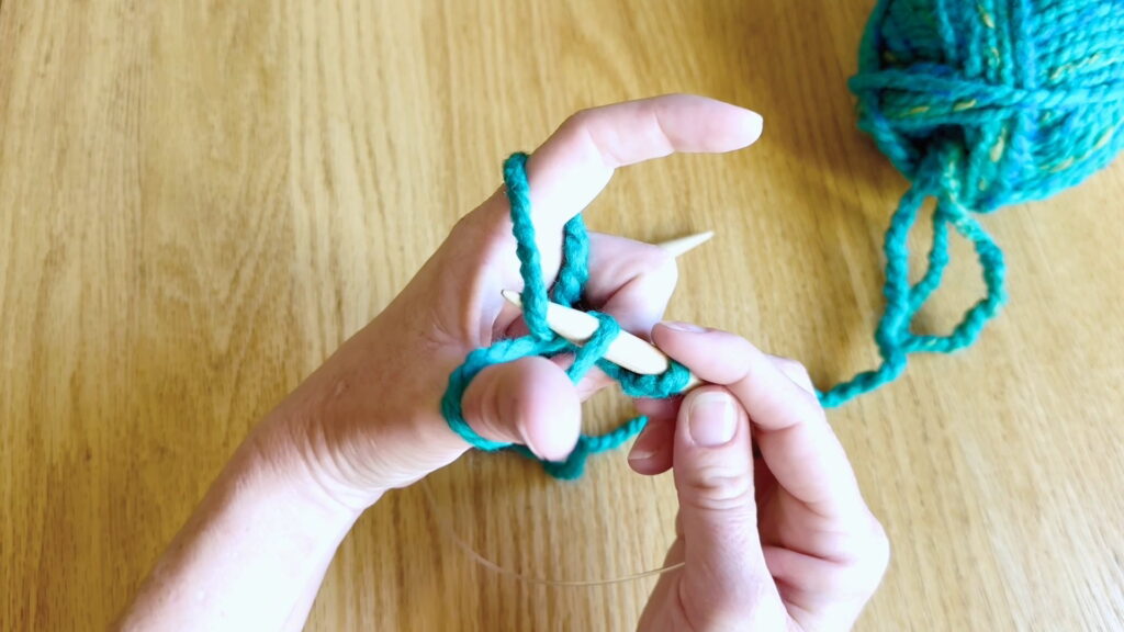 Needle tip grabbing the yarn strand and pulling it toward the thumb to form a new loop.