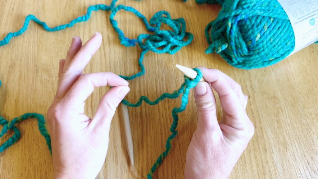 Knitting needle with a slipknot held in the right hand while the left thumb and pointer finger pinch together.