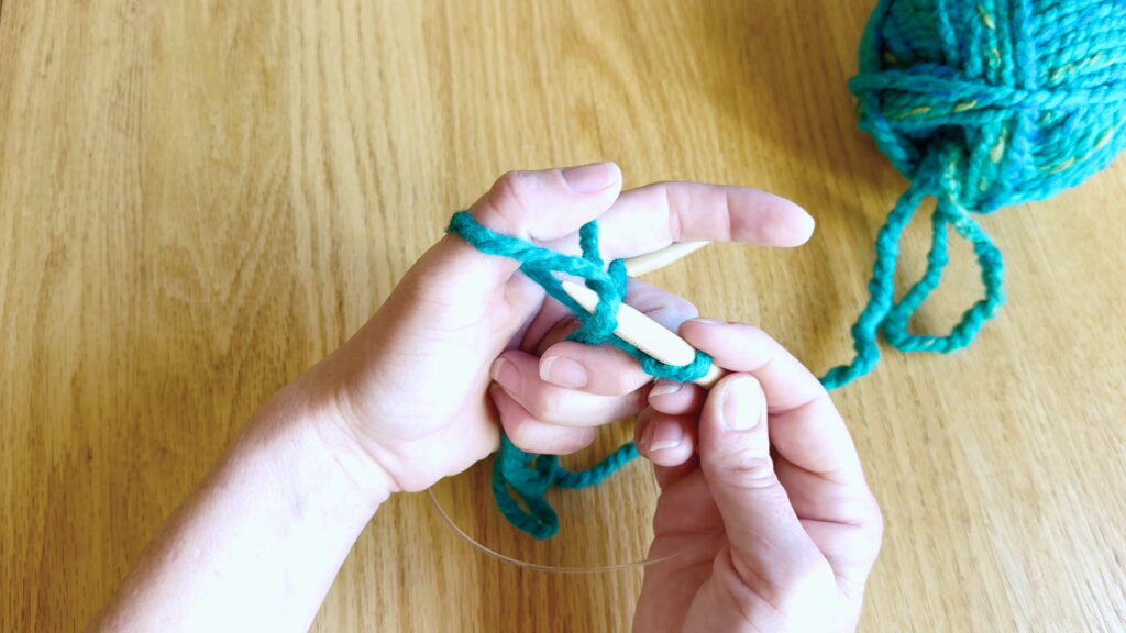 Needle tip pulling the yarn loop through the loop held on the thumb.