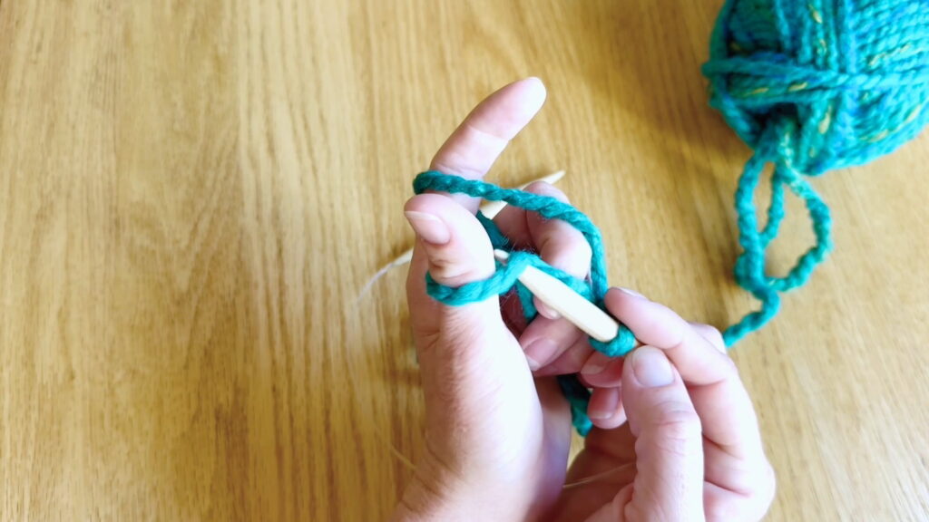 Knitting needle tip moving under the yarn strand looped around the left thumb.