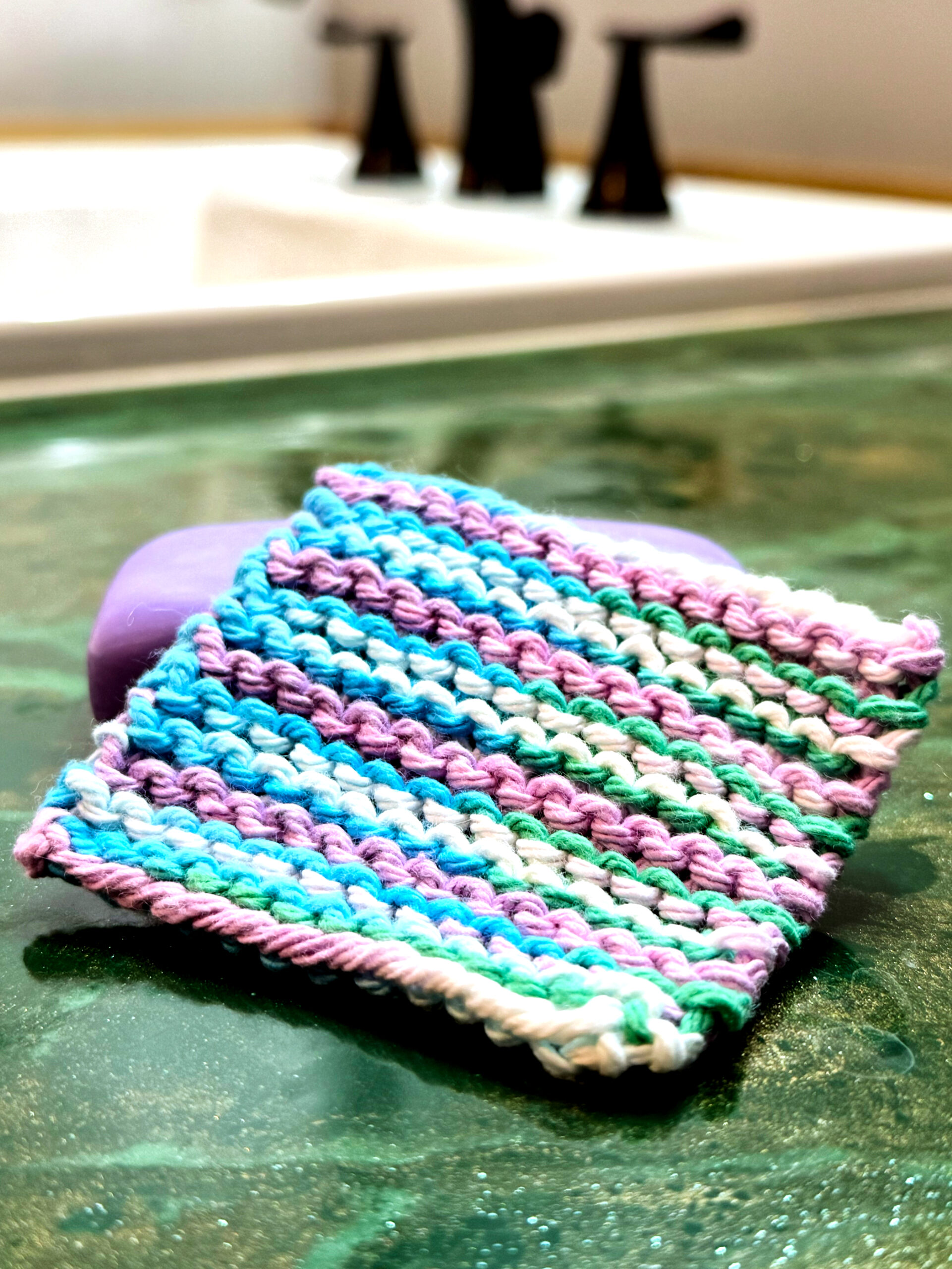 A completed knit mini washcloth near a bathroom sink and a purple bar of soap.