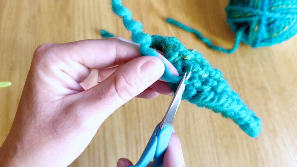 Cutting excess yarn near the secured knot after weaving in loose ends in beginner knitting.