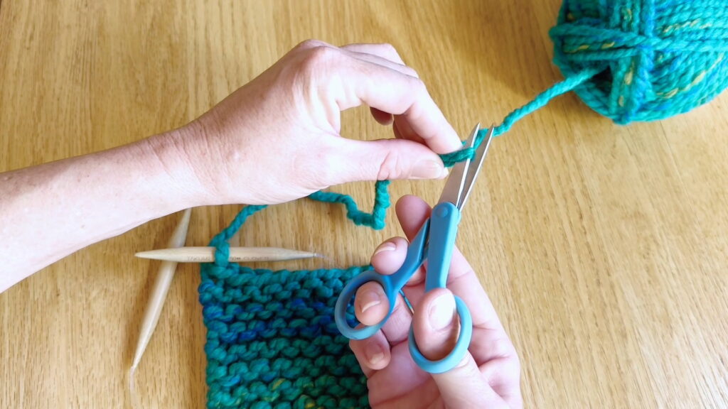 Measuring and cutting the yarn tail to finish a beginner knitting project after binding off.