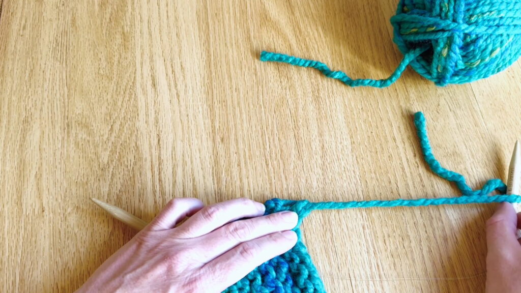 Pulling the yarn completely through the final loop to secure the end of the knitting.
