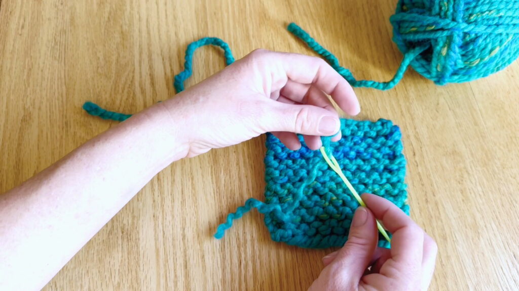 Threading half of the split yarn tail onto a darning needle to secure the loose end in beginner knitting.