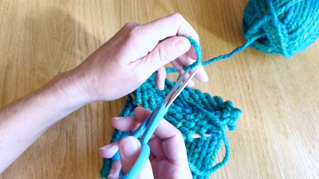Lesson 9, Kids' Knitting Guide – cutting the yarn tail to prepare for finishing a knitted hat.
