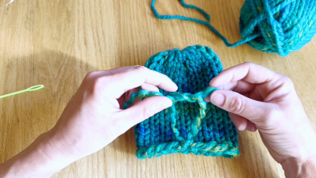 Lesson 10, Kids' Knitting Guide – tying the first knot to secure the bottom yarn tail of a knitted hat.