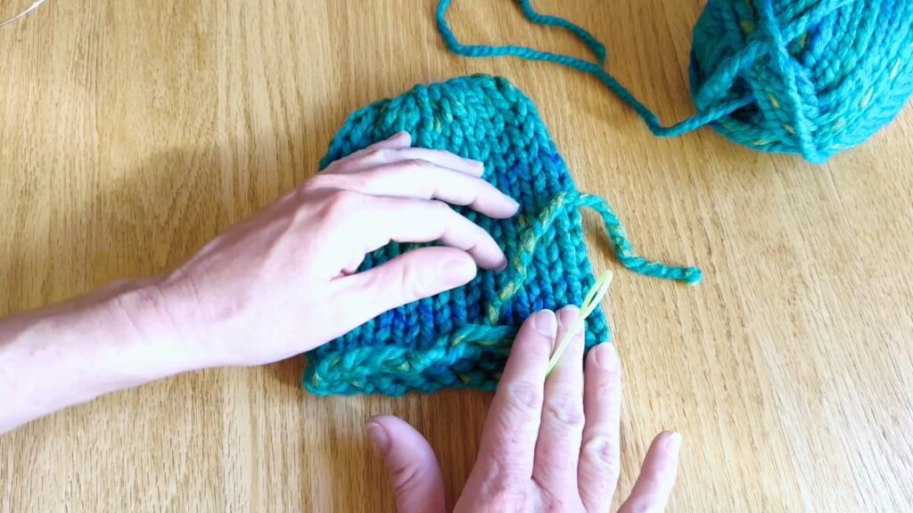 Lesson 10, Kids' Knitting Guide – preparing to weave in the remaining loose end at the bottom of a knitted hat.
