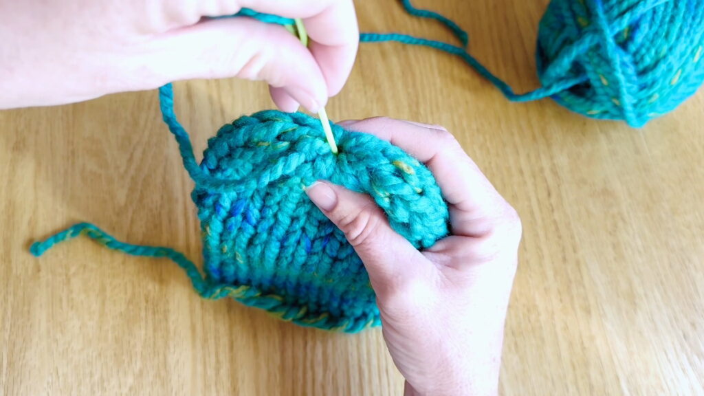 Lesson 10, Kids' Knitting Guide – inserting a darning needle into the top of a knitted hat to begin weaving in the yarn tail.