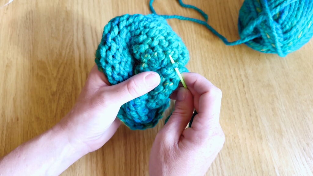Lesson 10, Kids' Knitting Guide – anchoring a yarn tail to a stitch near the top of a knitted hat.